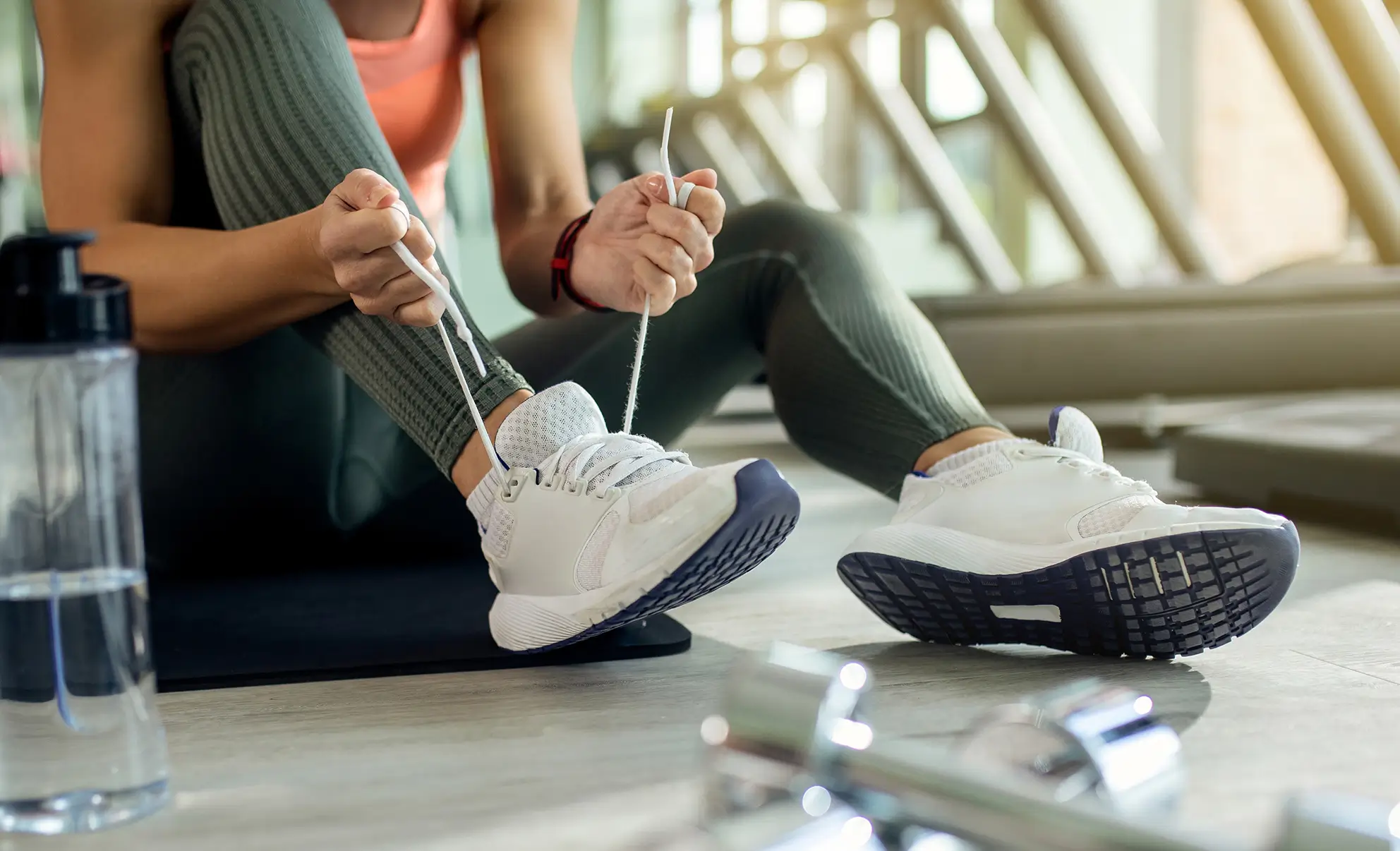 Person sitting on a gym mat tying white athletic shoes, with a water bottle and dumbbells on the floor nearby.