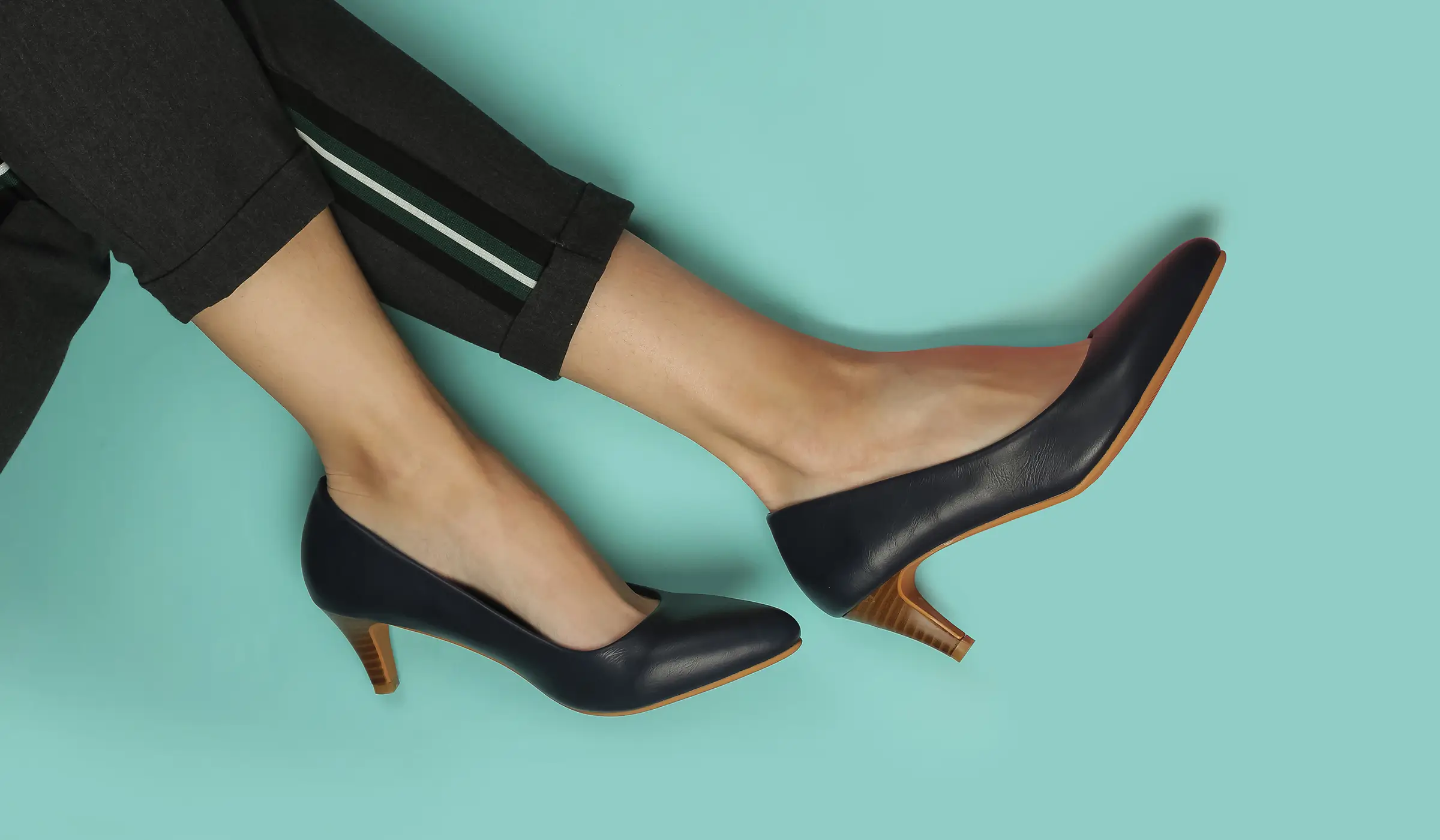 Person wearing black dress pants and navy blue low-heeled shoes with tan soles, legs extended against a light turquoise background.