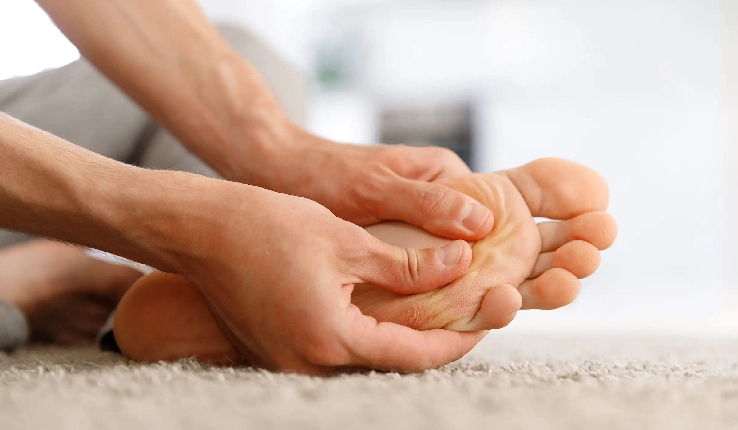 A person sits on a carpeted floor and massages the sole of their bare foot with both hands.