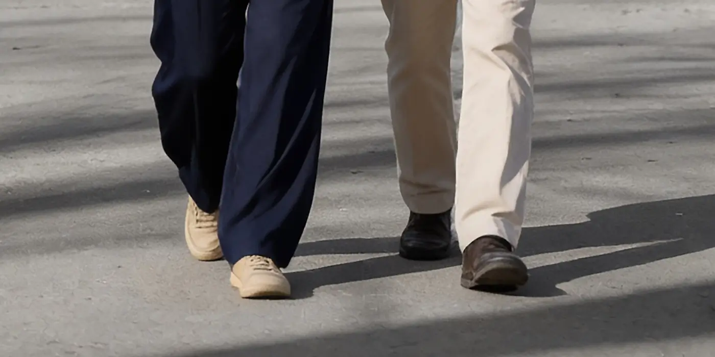 Two people walking side by side on a paved surface, one wearing dark blue pants and beige shoes, the other wearing light khaki pants and brown shoes.