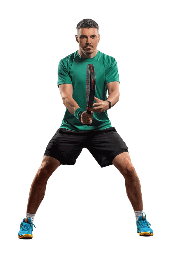 A man in a green shirt and black shorts stands in a ready position holding a tennis racket, preparing to play.